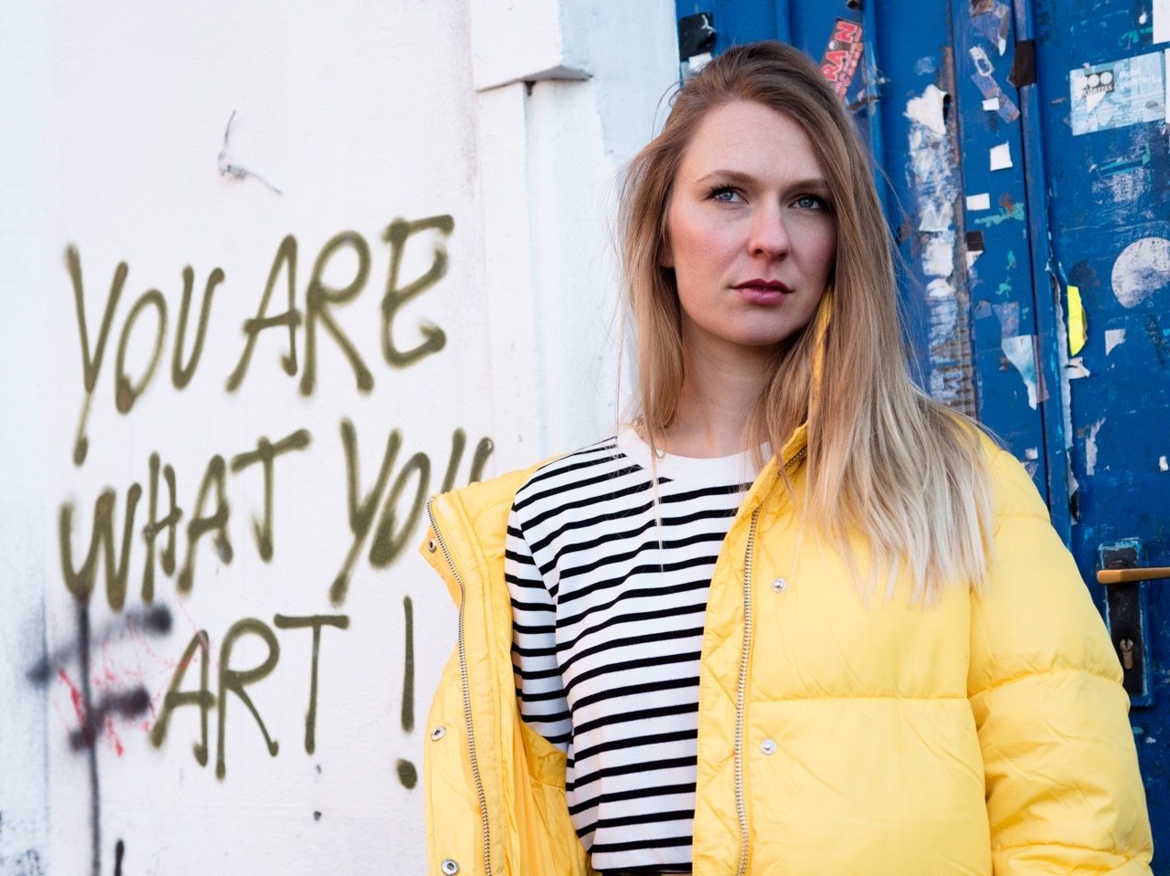 Street-Fashion-Porträt einer blonden Frau in gelber Steppjacke und gestreiftem Oberteil vor einer Graffitiwand