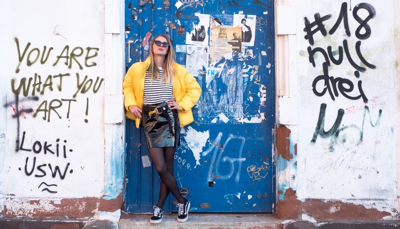 Ganzkörper-Modefoto einer Frau in gelber Jacke, Rock und Sneakern vor einer verwitterten, mit Graffiti bedeckten blauen Tür