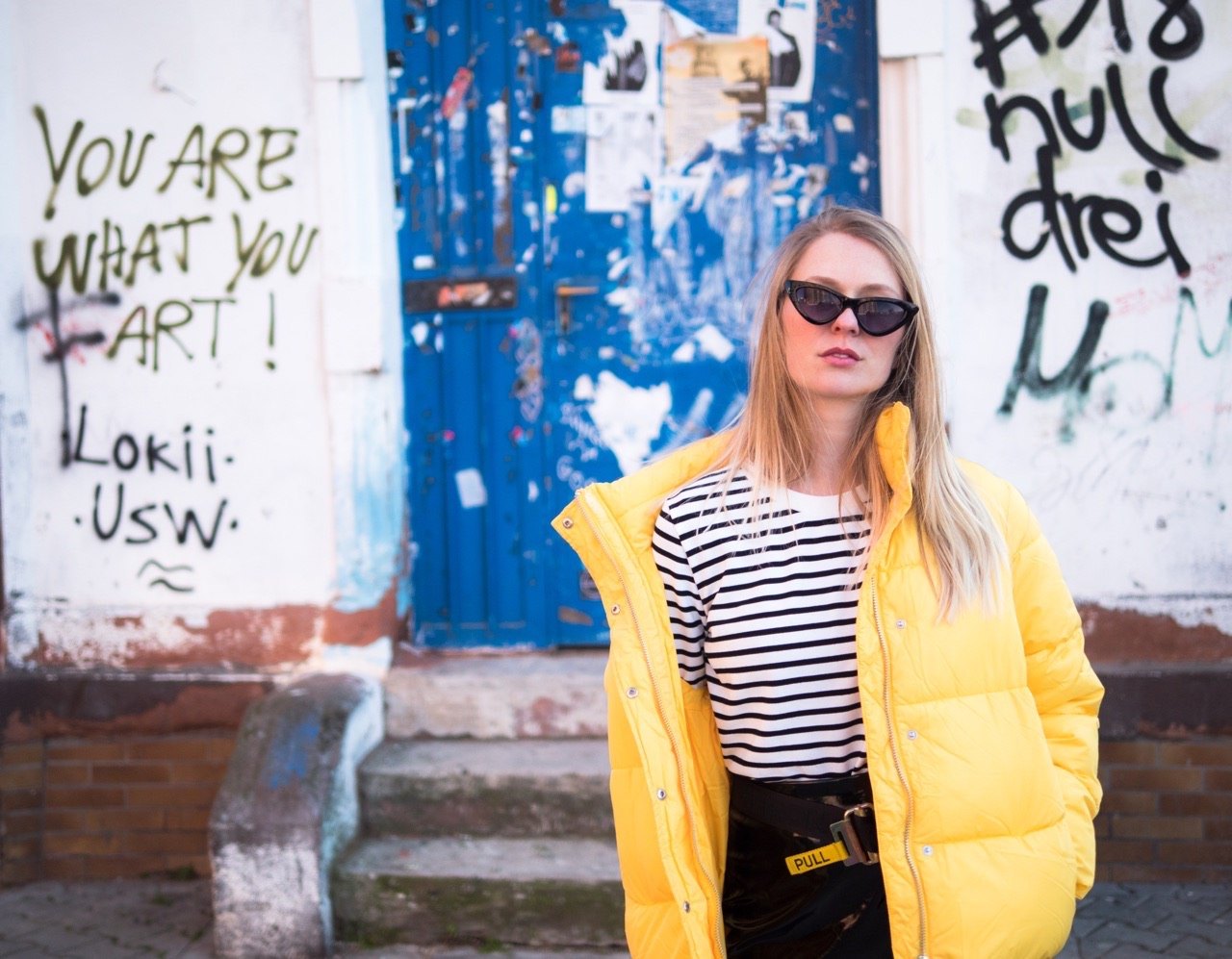 Street-Fashion-Foto einer Frau mit Sonnenbrille und gelber Steppjacke neben einer blauen Tür voller Graffiti