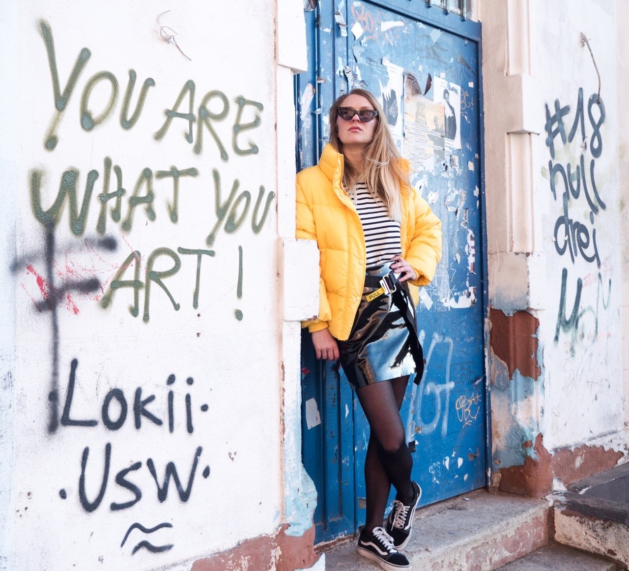 Street-Fashion-Shooting: Frau in gelber Steppjacke, gestreiftem Oberteil und Sonnenbrille an einer blauen Tür neben dem Graffiti 'You are what you art'