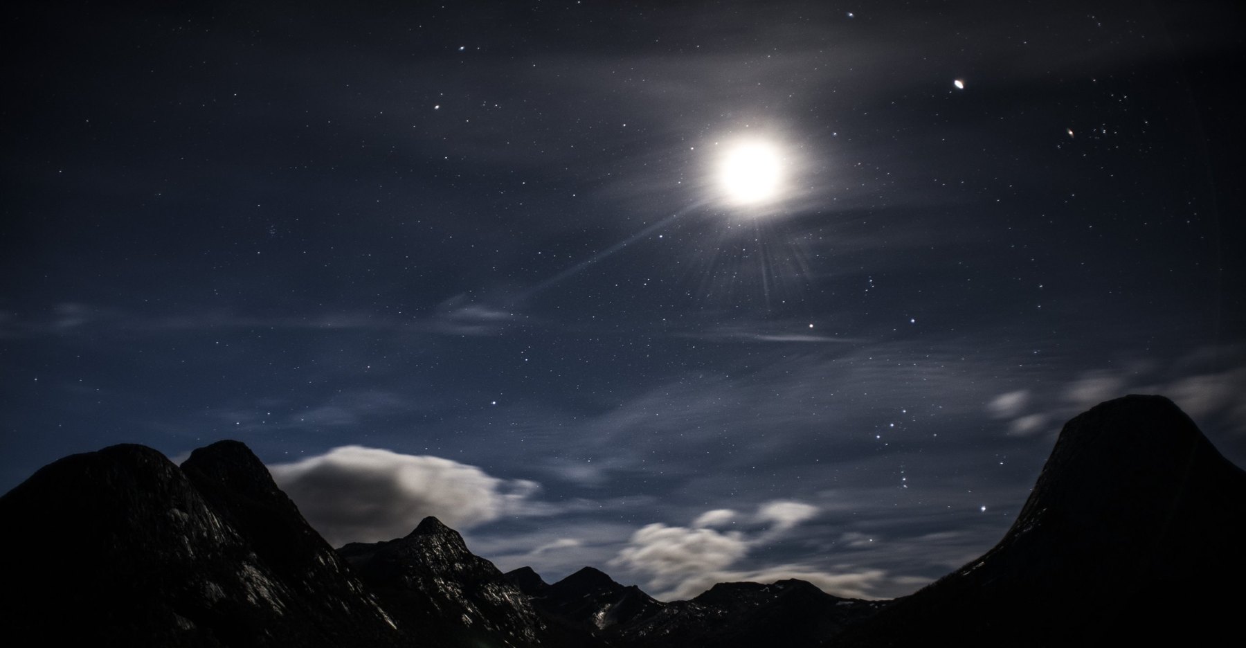 Nachtlandschaft: heller Mond und Sterne über dunklen Bergsilhouetten und tief hängenden Wolken