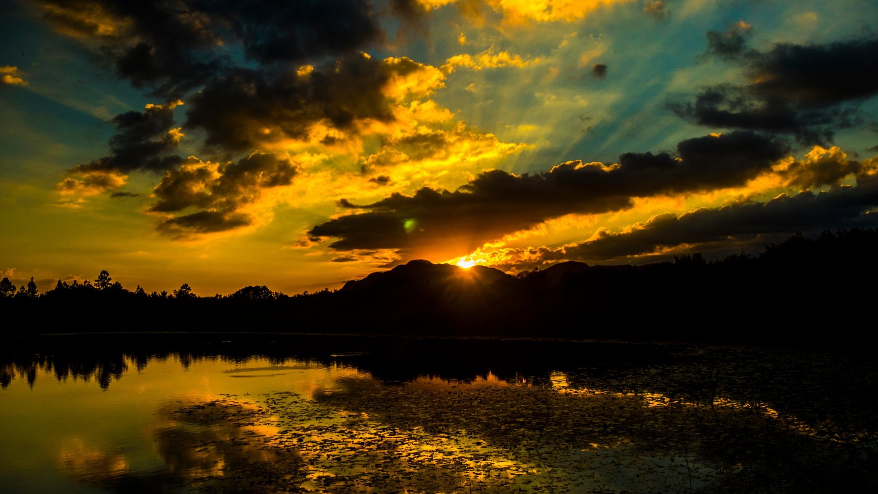 Dramatischer goldener Sonnenuntergang, der durch dunkle Wolken bricht und sich in einem stillen See spiegelt