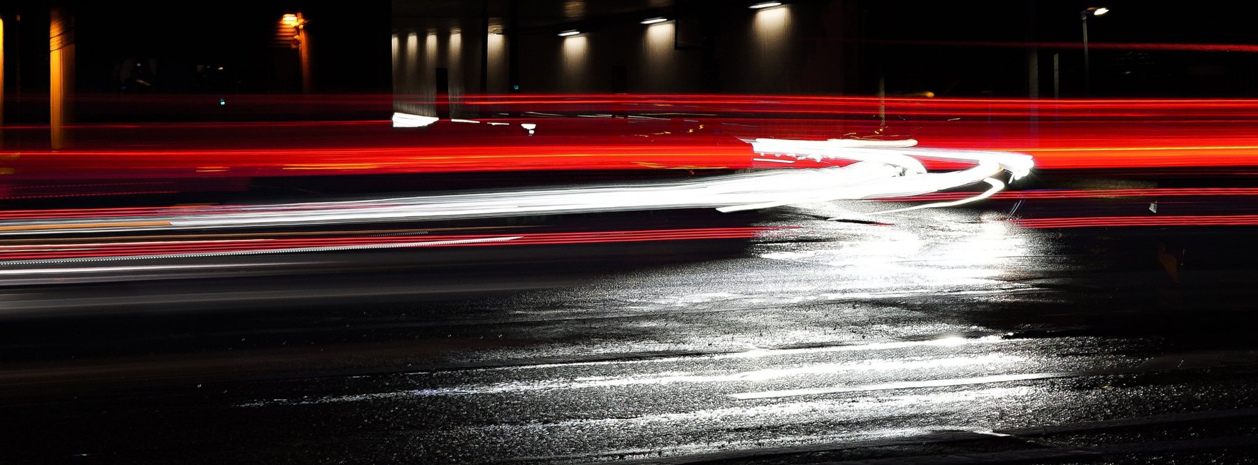 Nachtaufnahme mit Langzeitbelichtung: rote und weiße Lichtspuren von Autos auf einer nassen Stadtstraße