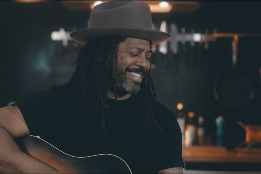 Lächelnder Musiker mit Dreadlocks und Filzhut spielt Akustikgitarre in einer schummrigen Bar