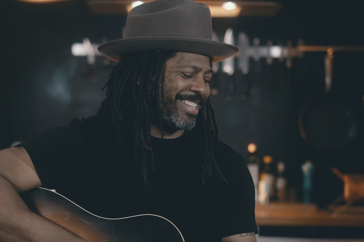 Lächelnder Musiker mit Dreadlocks und Filzhut spielt Akustikgitarre in einer schummrigen Bar