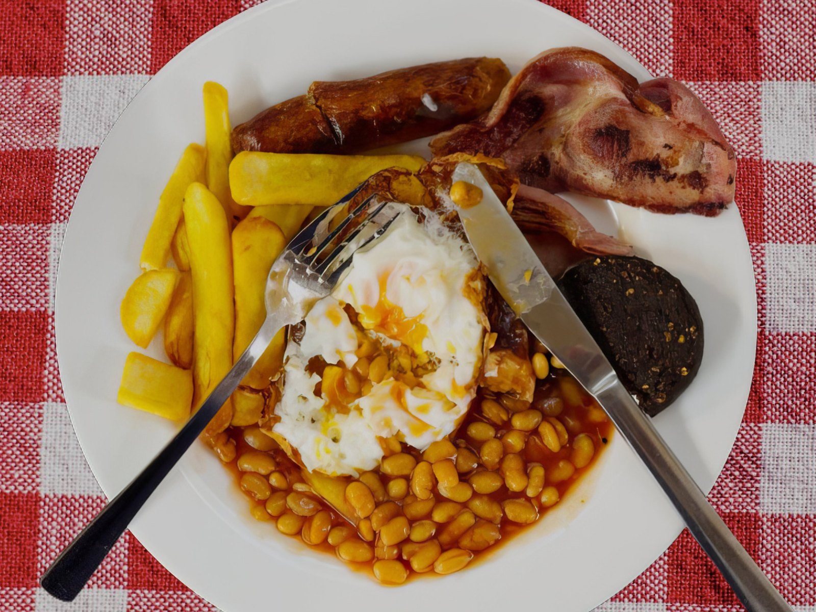 The English: ein Full English Breakfast mit Ei, Bohnen, Speck, Wurst, Black Pudding und Pommes auf einer rot karierten Tischdecke