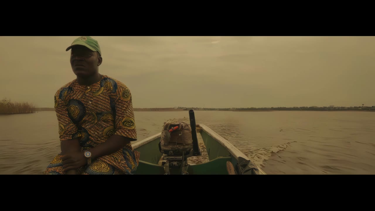 Trailer zu Dying to Clothe You: ein Mann in gemustertem Hemd steuert ein Boot über einen Fluss